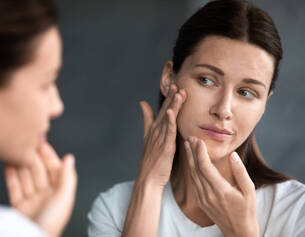 Una donna si guarda allo specchio mentre si prende cura della pelle del viso, mostrando espressione di attenzione.