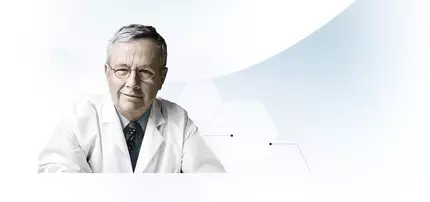 Un medico anziano con occhiali, vestito con un camice bianco, sorride guardando verso la camera.