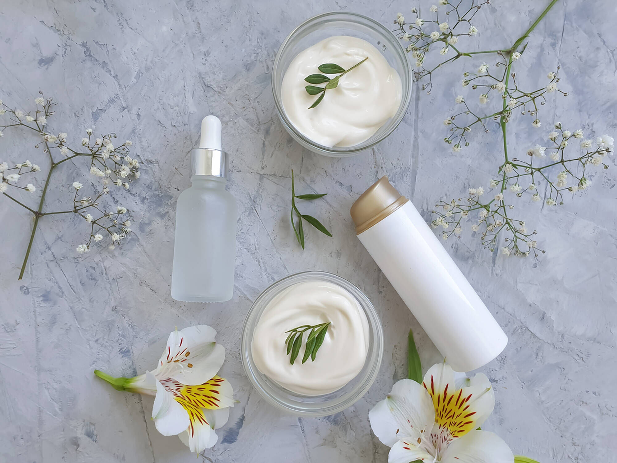 Creme per viso in barattoli con fiori e foglie, accompagnate da un flacone di siero trasparente su sfondo grigio.