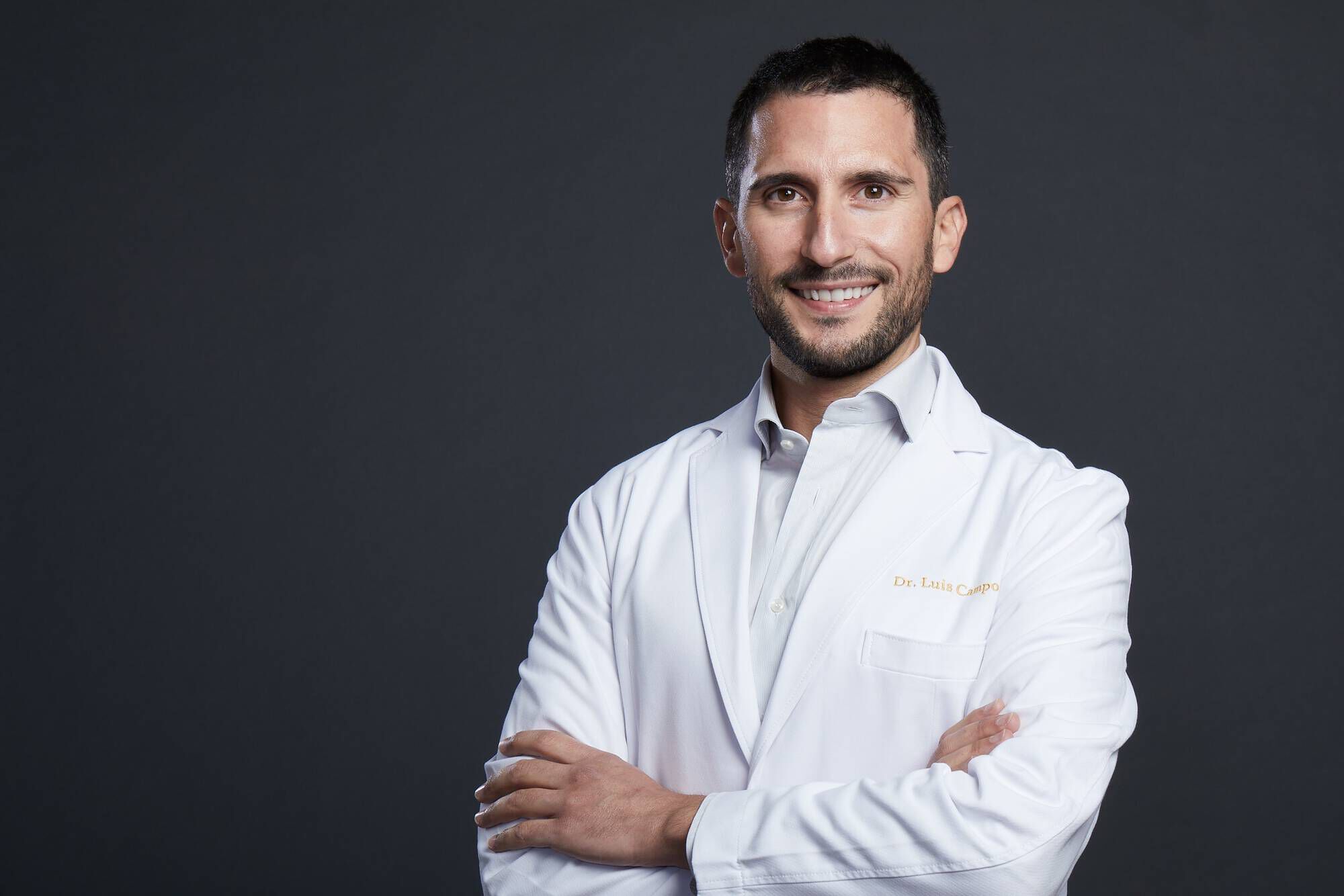 Un medico sorridente indossa un camice bianco, con le braccia incrociate, su uno sfondo scuro.