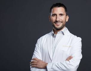 Un medico sorridente indossa un camice bianco, con le braccia incrociate, su uno sfondo scuro.