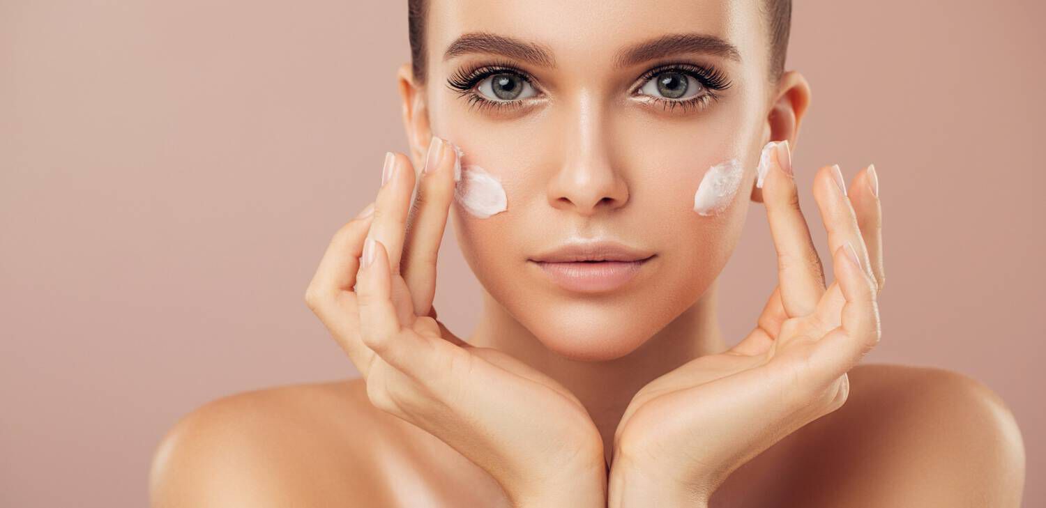 Giovane donna applica crema viso sul volto, sorridendo con occhi espressivi, su sfondo neutro.
