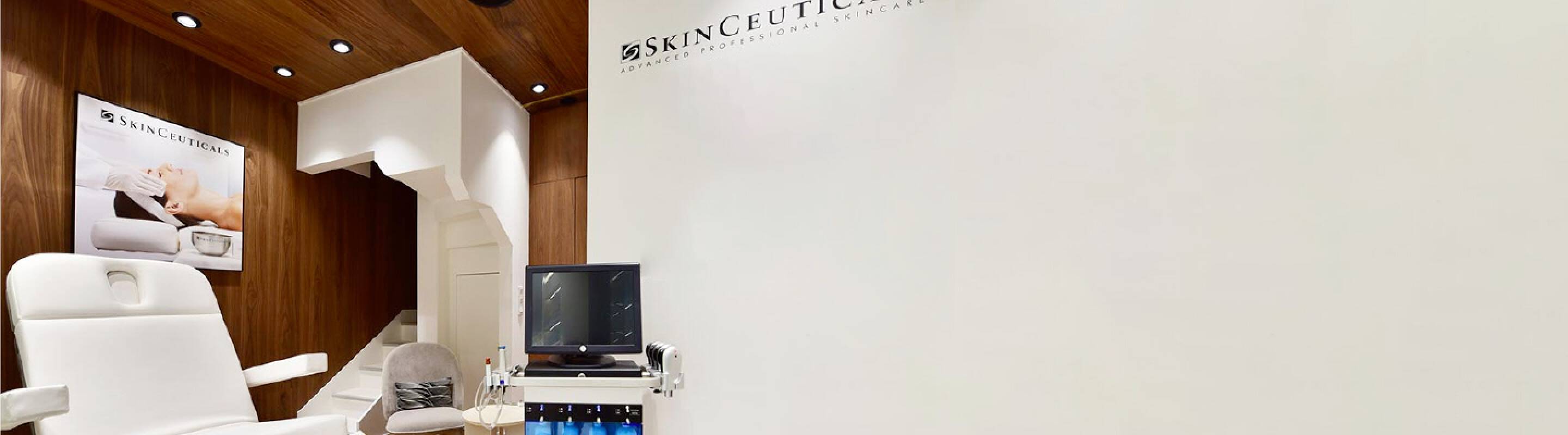 Alt text: Interno di uno studio dermatologico moderno, con pareti chiare e un lettino terapeutico. Sullo sfondo si vede un monitor e una presentazione del marchio Skinceuticals.