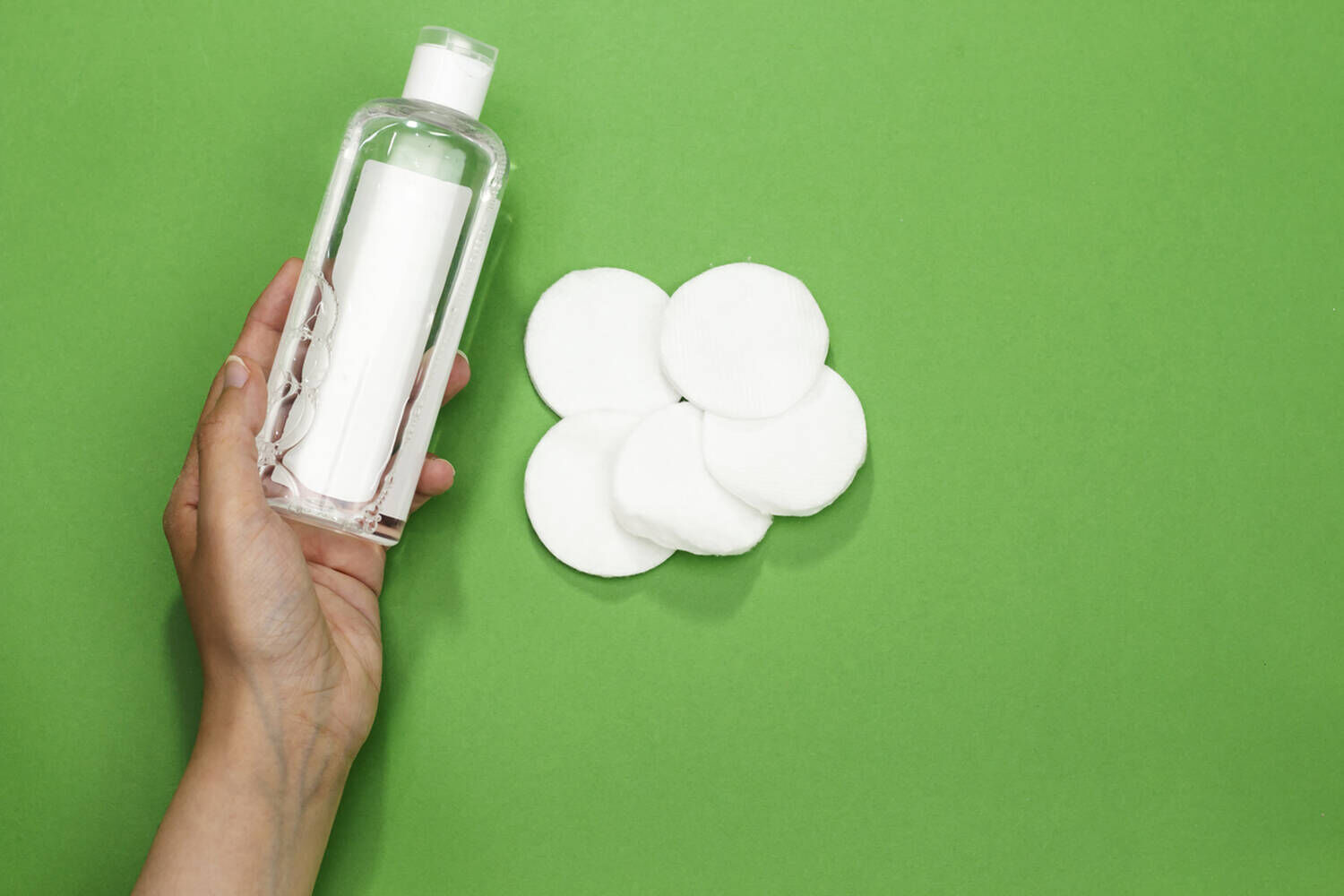 Una mano tiene una bottiglia di tonico accanto a dischetti di cotone su sfondo verde.