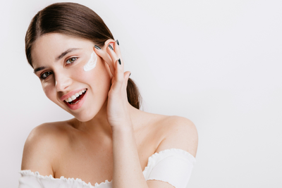 Una donna sorridente si applica crema bianca sulla guancia con il dito.