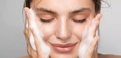 Una giovane donna che si lava il viso con un prodotto schiumoso, sorridendo e con gli occhi chiusi.