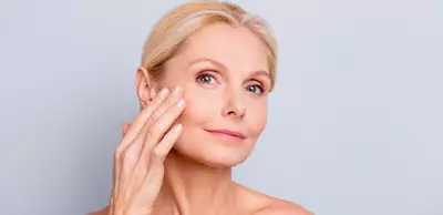 Una donna con i capelli biondi e lisci tocca delicatamente il suo viso sorridendo, davanti a uno sfondo chiaro.
