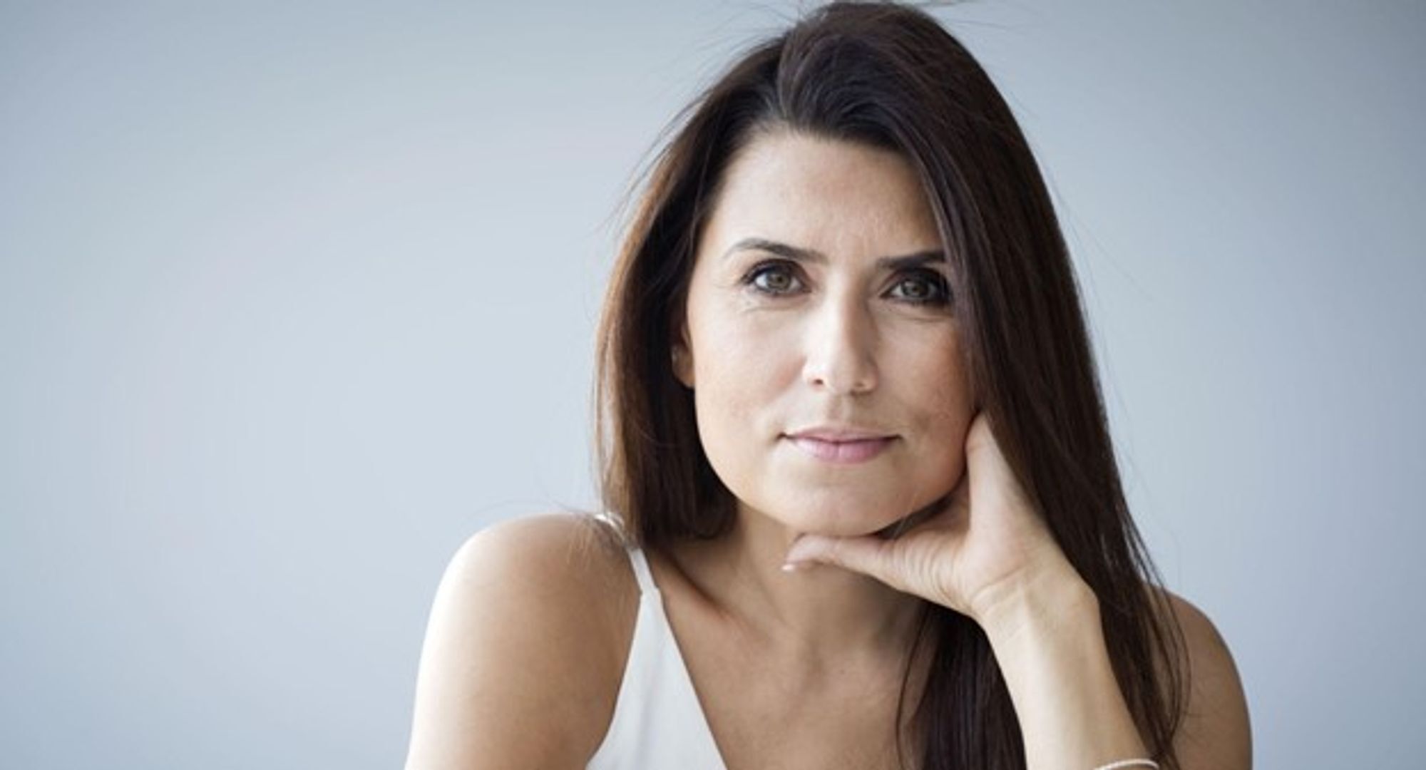 Ritratto di una donna con capelli lunghi e scuri, che guarda con espressione seria. Indossa una maglietta bianca e si appoggia con una mano sul viso. Sullo sfondo, una parete grigia chiara.