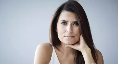 Ritratto di una donna con capelli lunghi e scuri, che guarda con espressione seria. Indossa una maglietta bianca e si appoggia con una mano sul viso. Sullo sfondo, una parete grigia chiara.