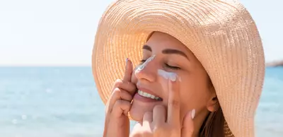 Una giovane donna con un cappello di paglia applica la crema solare sul viso, sorridendo mentre si trova sulla spiaggia. Sullo sfondo si vede il mare.