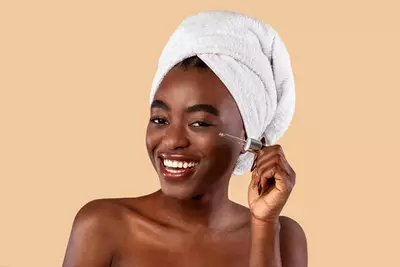 Una donna sorridente con un asciugamano bianco in testa applica un siero per la cura della pelle.