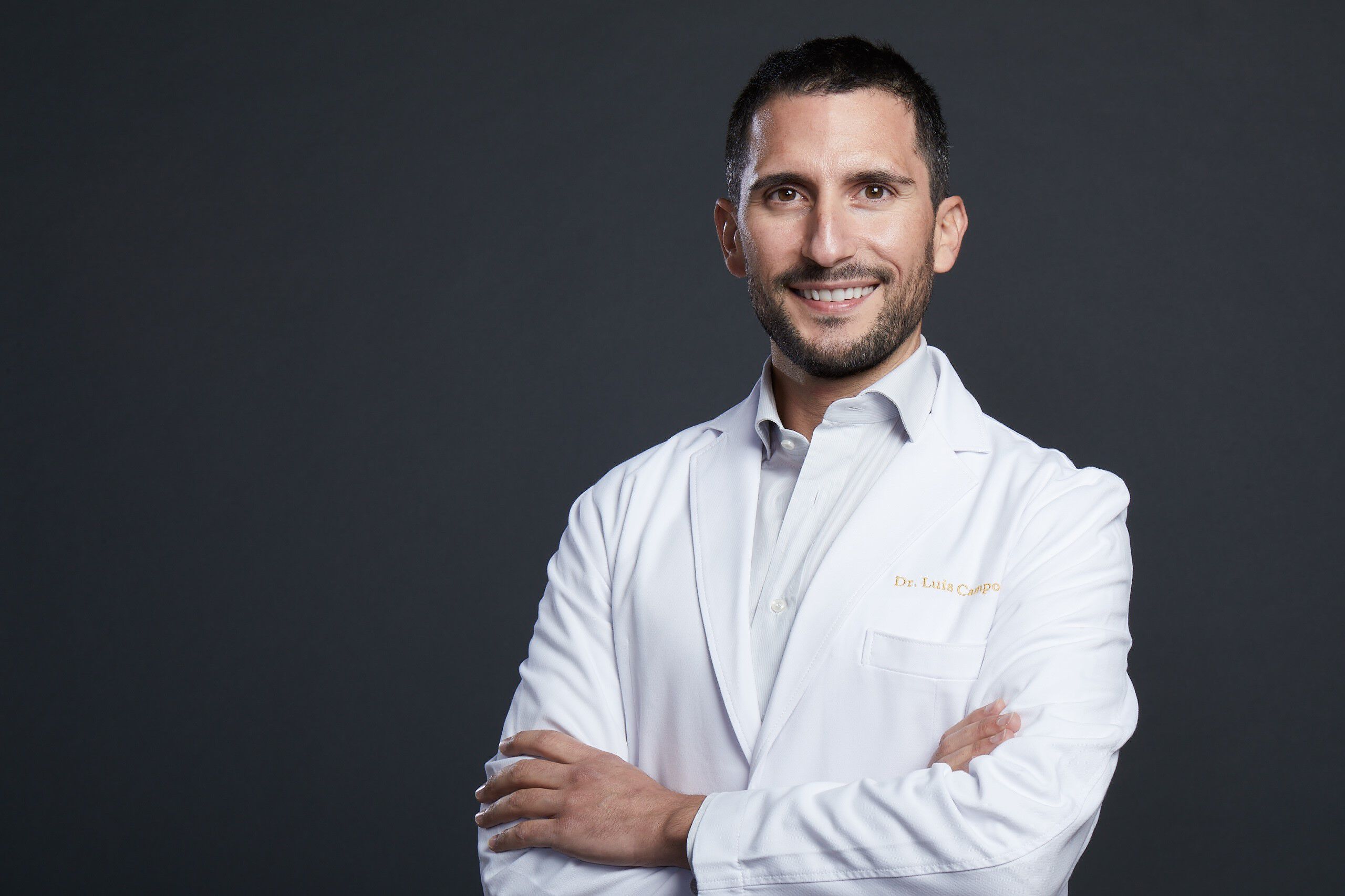 Un medico in camice bianco sorride, con le braccia incrociate, su uno sfondo scuro.